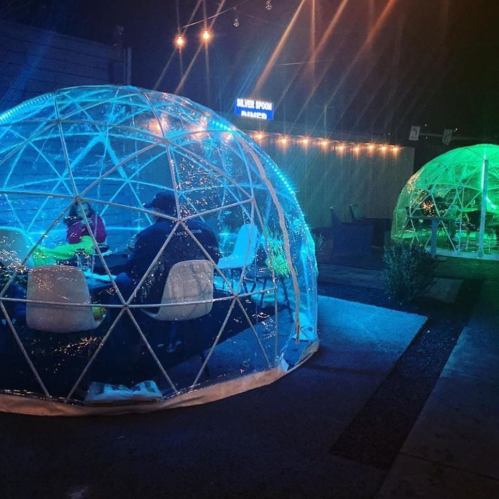 Two illuminated igloo-like structures with people dining inside, surrounded by string lights in a nighttime setting.