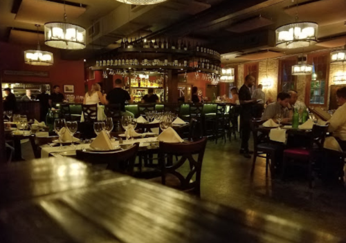 A dimly lit restaurant with tables set for dining, a bar in the background, and patrons enjoying their meals.