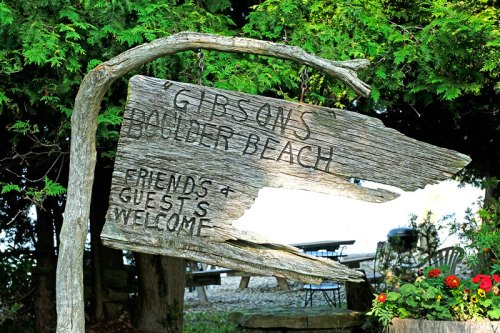 Weathered wooden sign reading "Gibson's Ponder Beach" with "Friends & Guests Welcome" surrounded by greenery.