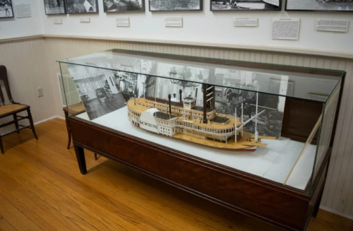 A detailed model of a steamboat displayed in a glass case, with historical photos on the wall behind it.