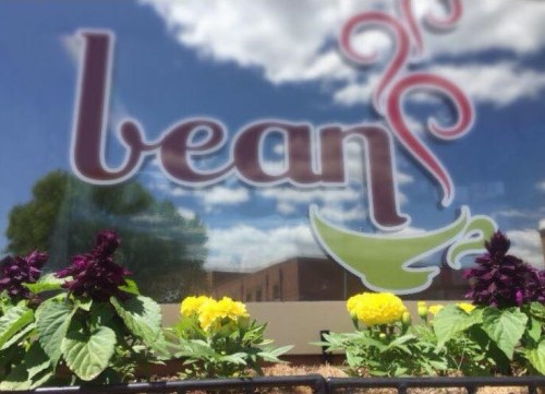 A window with the word "bean" and colorful flowers in front, reflecting a blue sky with clouds.