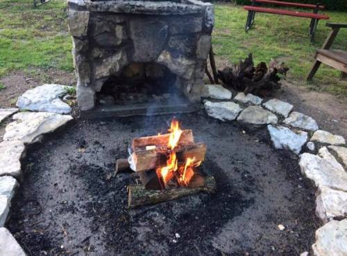 A stone fire pit with a roaring fire surrounded by rocks and grass, with benches in the background.