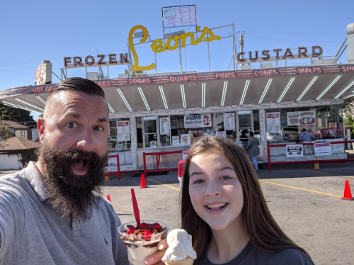 A man and a girl smile in front of Leon's Frozen Custard, holding ice cream treats on a sunny day.