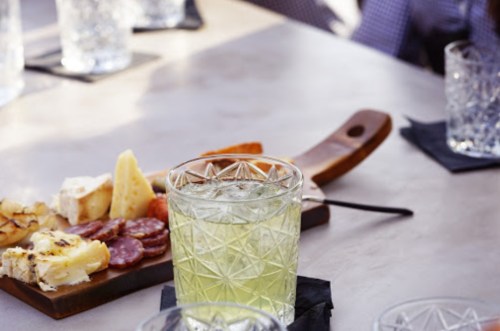 A close-up of a cocktail glass on a table with a charcuterie board featuring cheeses and meats in the background.