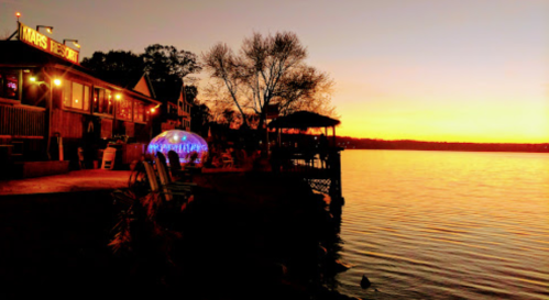 Sunset over a lake with a restaurant and illuminated dome seating by the water's edge. Trees silhouette against the sky.