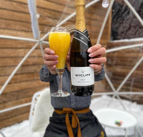 A person holds a glass of orange mimosa and a bottle of Wycliff sparkling wine in a cozy setting.