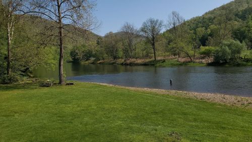 A serene river scene with lush green grass, trees, and a person fishing in the water under a clear blue sky.