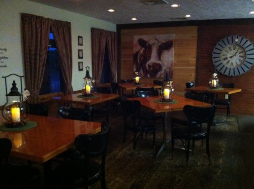 Cozy restaurant interior with wooden tables, black chairs, lanterns, and a cow painting on the wall. Warm, inviting atmosphere.