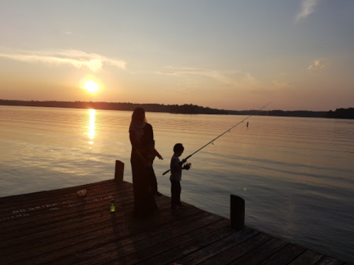 A woman and a child fishing from a wooden dock at sunset, with calm water reflecting the colorful sky.