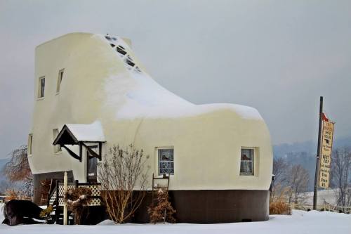 A whimsical shoe-shaped house covered in snow, with a sign nearby and a cloudy sky in the background.