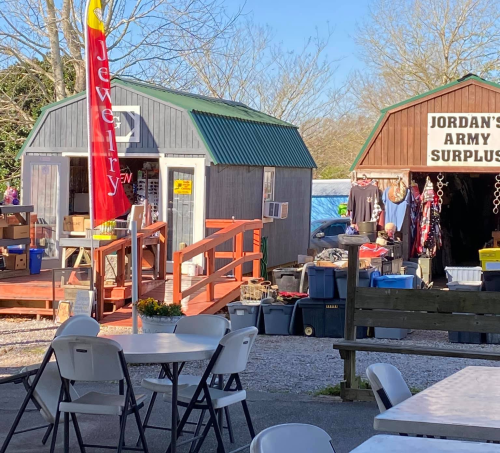 A sunny outdoor market with two storage sheds, a jewelry sign, and tables set up for visitors.