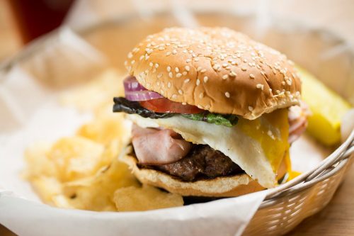 A close-up of a sesame seed burger with lettuce, tomato, onion, ham, and a fried egg, served with potato chips.