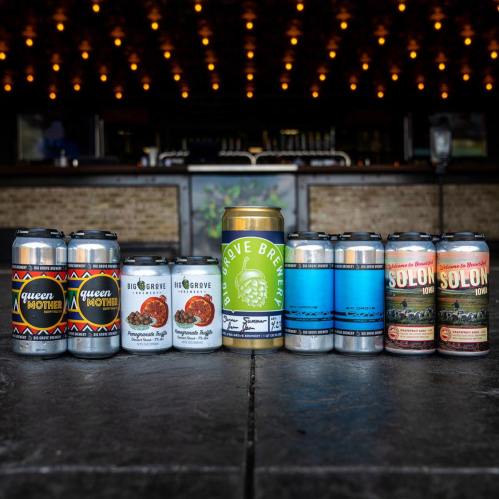 A variety of craft beer cans displayed on a table, with a bar backdrop and warm lighting in the background.