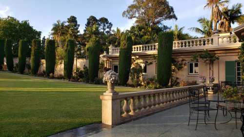 A landscaped garden with tall trees, a stone railing, and a patio area beside a beautiful house.