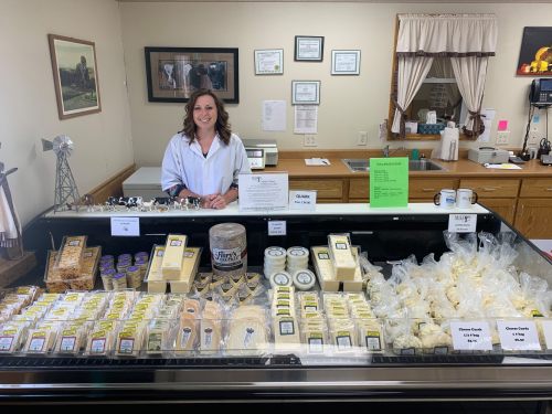A smiling woman in a white coat stands behind a display of various cheeses and dairy products in a shop.