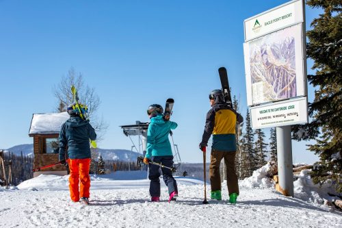 Three skiers in colorful gear walk towards a ski lift sign in a snowy landscape, ready for a day on the slopes.
