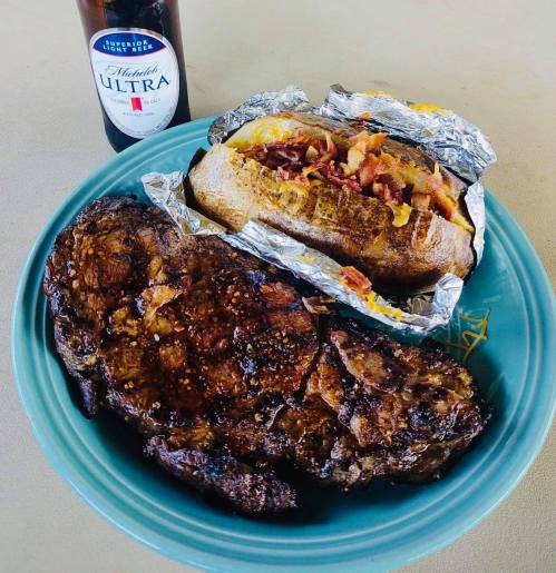 A plate with a grilled steak, a loaded baked potato wrapped in foil, and a bottle of Michelob Ultra beer.