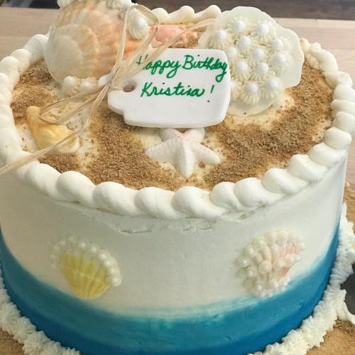A decorated birthday cake with seashells, sand, and a tag that says "Happy Birthday Kristia!" on top.