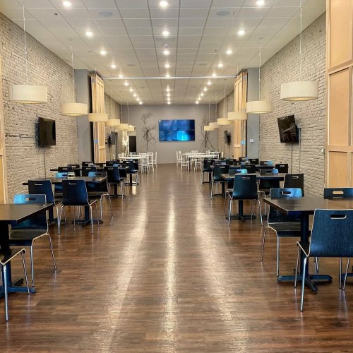 A spacious dining area with wooden floors, black tables, and chairs, featuring pendant lights and wall-mounted TVs.