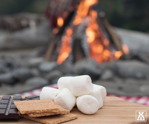 A wooden board with graham crackers, marshmallows, and chocolate in front of a campfire.