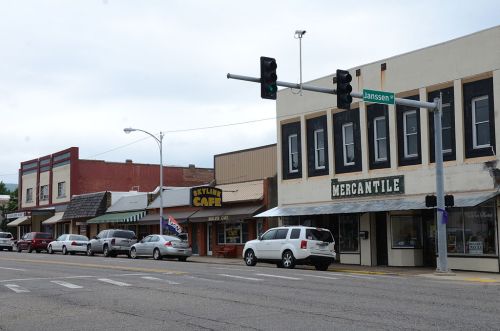 A street view of a small town featuring shops, a café, and traffic lights under a cloudy sky.