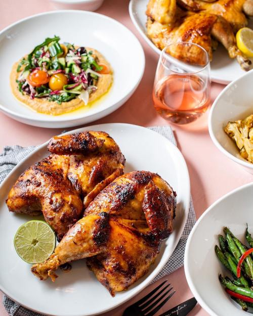 A plate of roasted chicken with lime, surrounded by salads and a glass of rosé wine on a pink tablecloth.