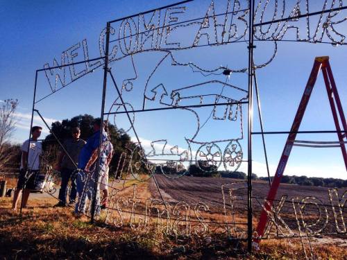Three people stand near a large wire frame sign that reads "Welcome Aboard" with a train design, set in a field.