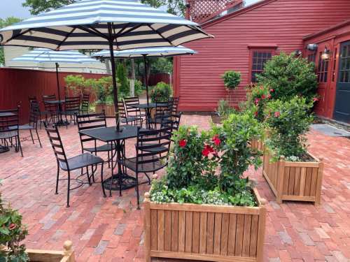 Outdoor patio with black tables and chairs, striped umbrellas, and flower planters against a red building.