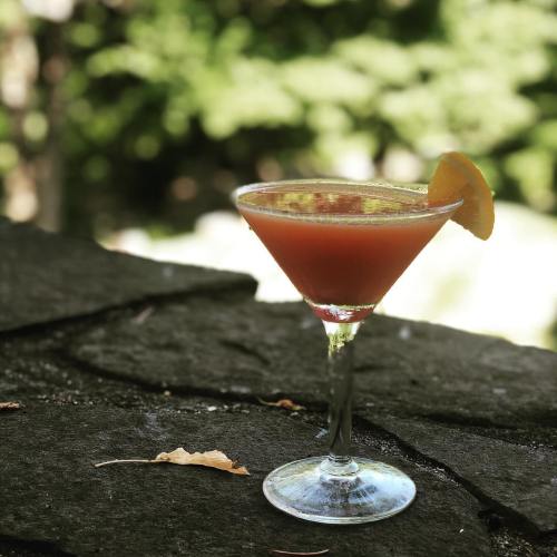 A vibrant cocktail in a martini glass, garnished with an orange slice, sits on a stone surface with a blurred green background.