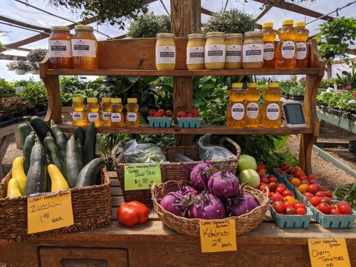 A wooden stand displays jars of honey and fresh vegetables, including zucchini, kohlrabi, and cherry tomatoes.