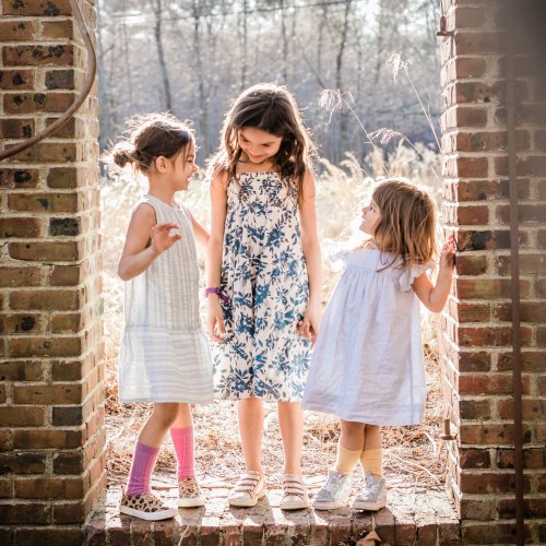 Three young girls stand together in a sunlit doorway, smiling and holding hands, surrounded by nature.