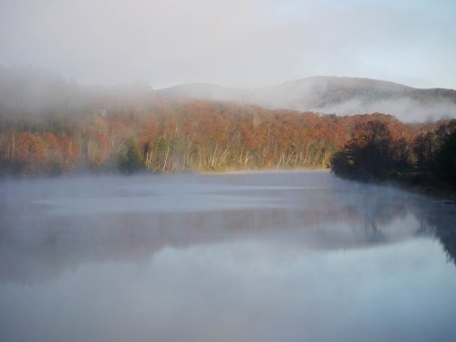 A serene lake surrounded by mist and autumn-colored trees, reflecting the tranquil landscape.