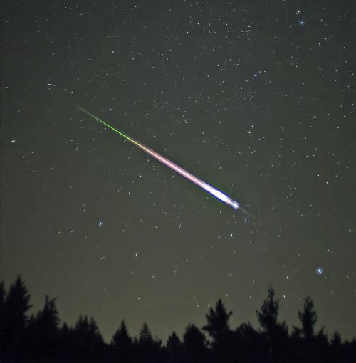 A bright meteor streaks across a starry night sky, with colorful trails above a silhouette of trees.