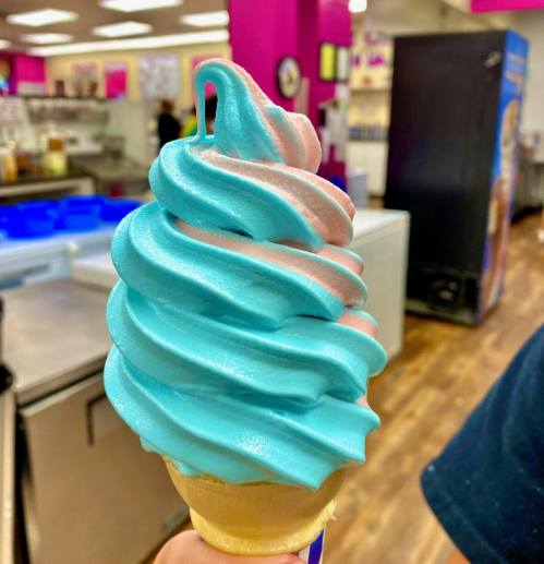 A close-up of a soft serve ice cream cone with blue and pink swirled toppings.