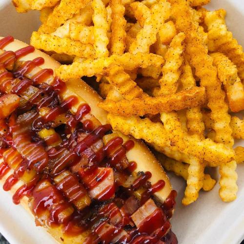A hot dog topped with barbecue sauce and diced toppings, served with a side of crispy seasoned crinkle-cut fries.