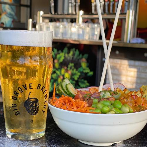 A glass of beer next to a bowl of colorful noodles and vegetables, with chopsticks resting on the bowl.