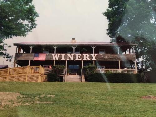 A rustic winery building with a large American flag, surrounded by green grass and trees.