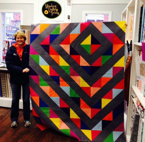 A woman stands beside a colorful geometric quilt displayed in a quilting shop, surrounded by fabric shelves.