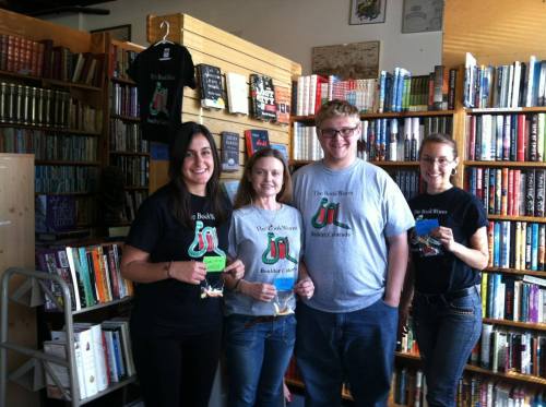 Four people stand in a bookstore, holding bookmarks and wearing matching t-shirts with a book-themed design.