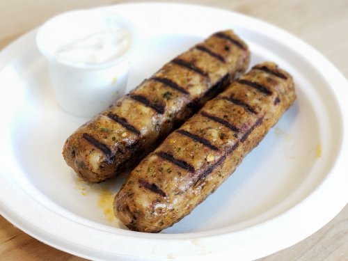 Two grilled kebabs on a paper plate, served with a small cup of white sauce.
