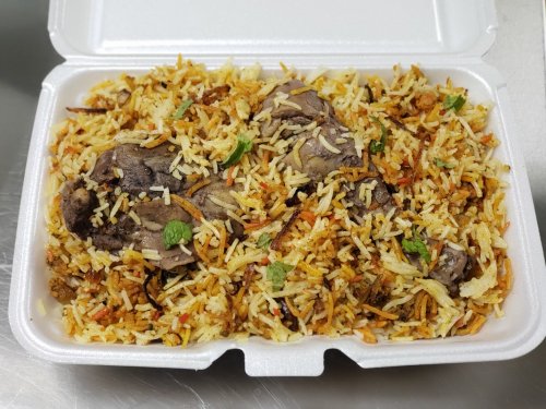 A takeout container filled with flavorful biryani rice topped with pieces of tender meat and garnished with herbs.