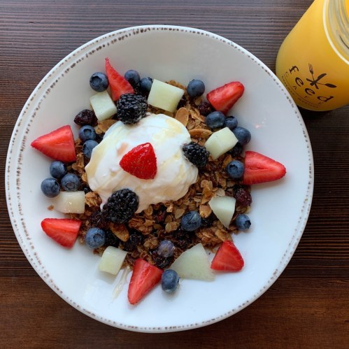 A bowl of granola topped with yogurt, strawberries, blueberries, blackberries, and melon, beside a bottle of juice.
