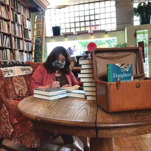 A woman in a mask sits at a wooden table, signing books in a cozy library filled with shelves and natural light.