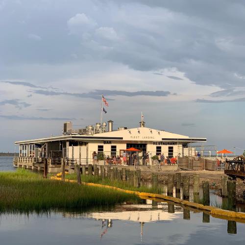A waterfront restaurant with a bar, surrounded by water and greenery, under a cloudy sky.
