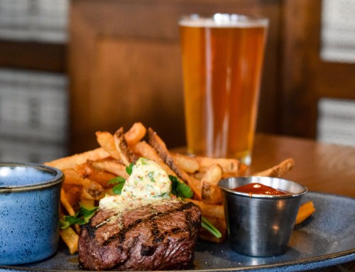 A plate with a grilled steak topped with herb butter, fries, and dipping sauces, alongside a glass of beer.