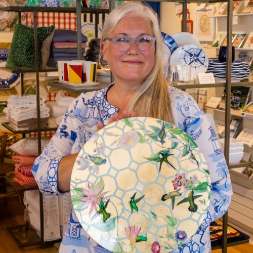 A woman with glasses holds a decorative plate featuring floral and hummingbird designs, smiling in a store.