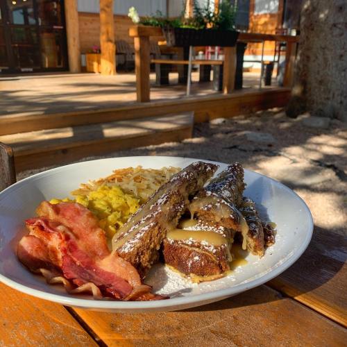 A plate of French toast with syrup, crispy bacon, scrambled eggs, and hash browns, set outdoors in a cozy setting.