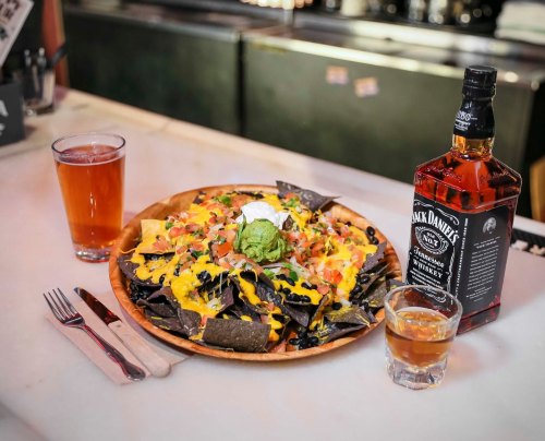 A wooden plate of nachos topped with cheese, guacamole, and sour cream, alongside drinks and a bottle of whiskey.