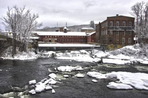 A snowy landscape featuring a river with ice, surrounded by historic brick buildings and bare trees.