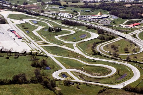 Aerial view of a winding racetrack surrounded by green fields and a parking area, with buildings in the background.
